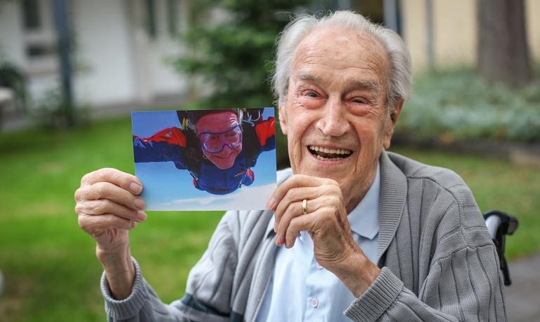 Ein Rentner zeigt ein Foto in die Kamera, das ihn beim Fallschirmsprung zeigt.